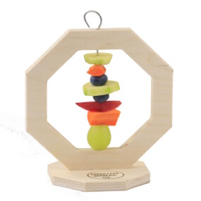 Das Foto zeigt einen Snackständer für Hamster aus Holz. Auf dem Spieß sind Trauben, Karotten und Heidelbeeren aufgespießt